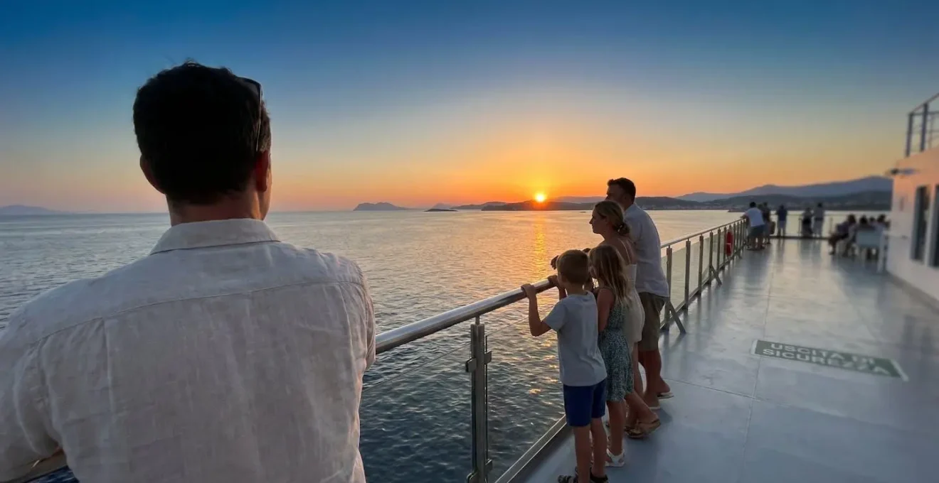 Una famiglia vista di spalle sulla prua di un traghetto moderno al tramonto, che osserva l'orizzonte del Mediterraneo con atmosfera serena