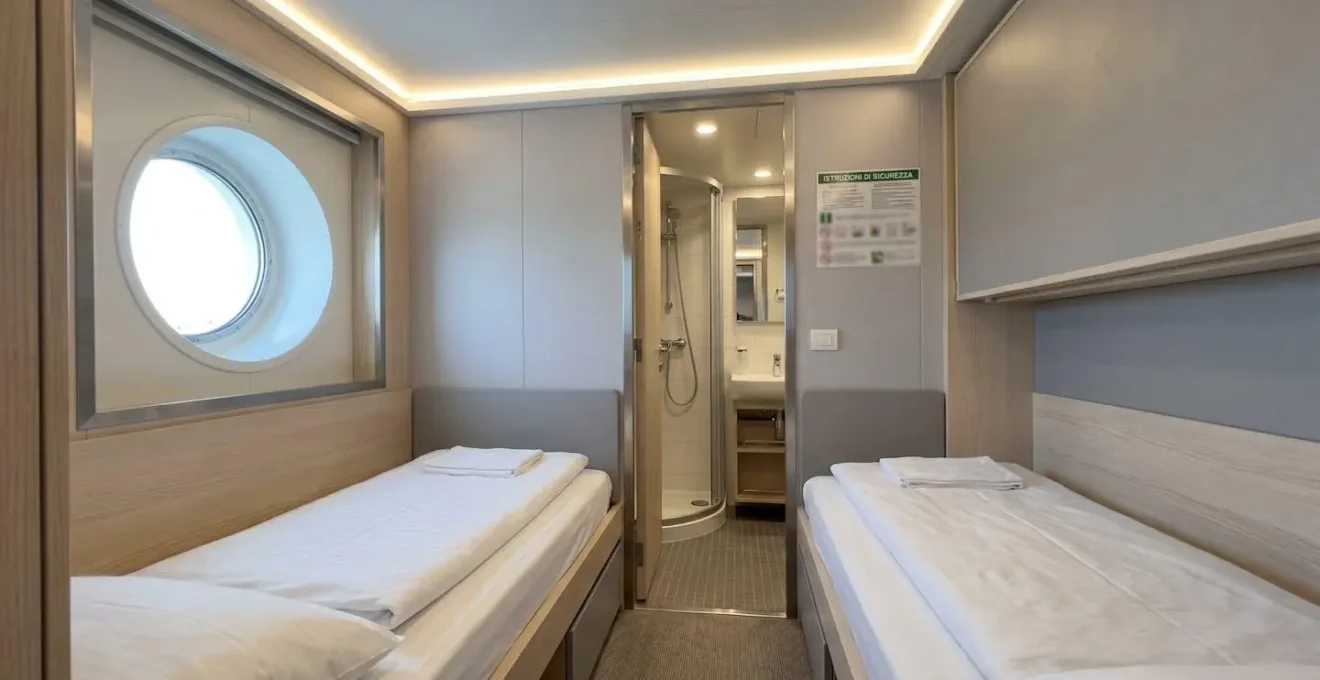 Interno luminoso di una cabina traghetto moderna con letti gemelli preparati, bagno privato visibile sullo sfondo e ambiente pulito e accogliente